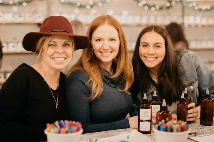 Three women smiling at a table with bottles and craft supplies.