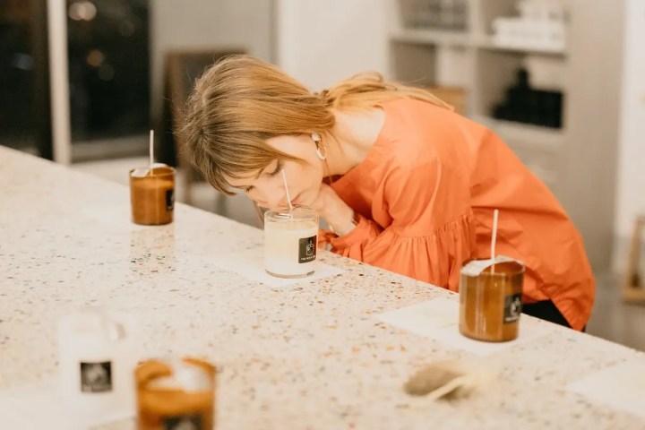 Woman in orange top smelling a candle in a bright room.
