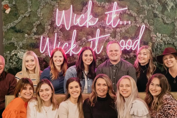 Group of people smiling in front of a neon sign on a green wall.