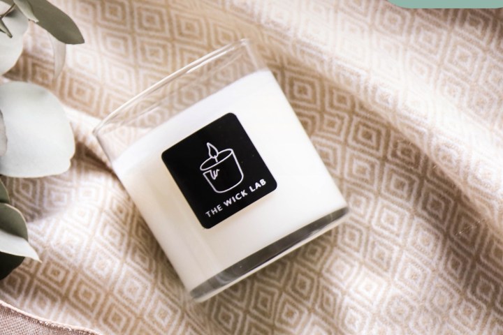 White candle in a clear glass on patterned fabric with greenery.